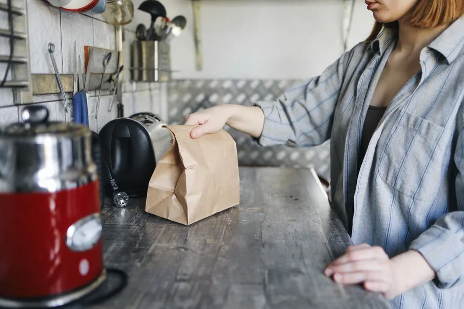 A person stands at a kitchen counter holding a closed brown paper bag. Several generic countertop appliances and kitchen utensils are visible, but no cooking ingredients are visibly identifiable.
