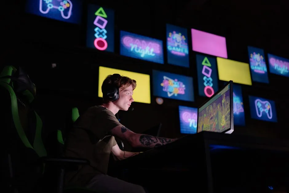 Side view of a headset-wearing player seated at a desktop monitor in a dark gaming venue, with colorful gaming-themed screens and controller-symbol graphics in the background. The monitor content is indistinct and does not clearly show digital-item listing rows or a comparison interface.