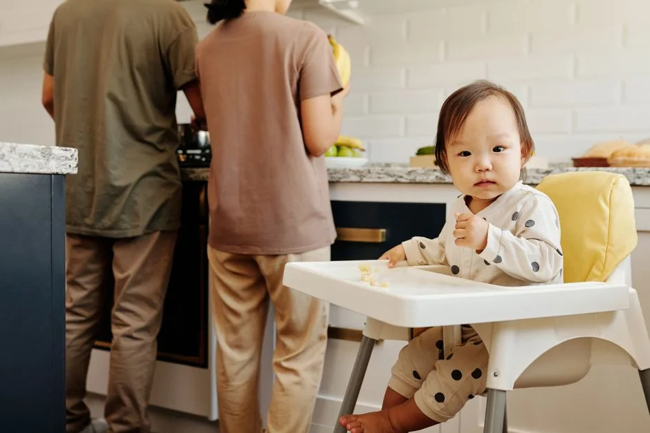 주방에서 두 성인이 조리대 앞에 서 있고, 유아가 흰색 식탁 의자에 앉아 있는 장면입니다. 젖병이나 여러 수유용품은 보이지 않습니다.