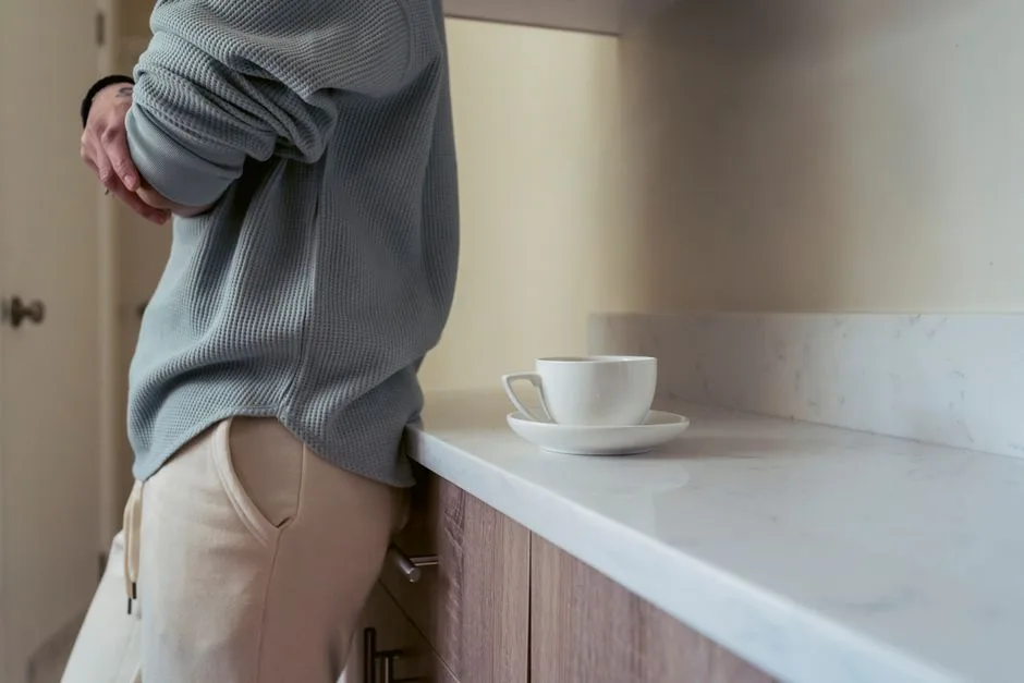 A person stands beside a kitchen countertop with one plain white cup on a saucer. No visible logos, brand text, watermarks, prices, promotions, ecommerce UI, reviews, or recognizable branded product designs appear.