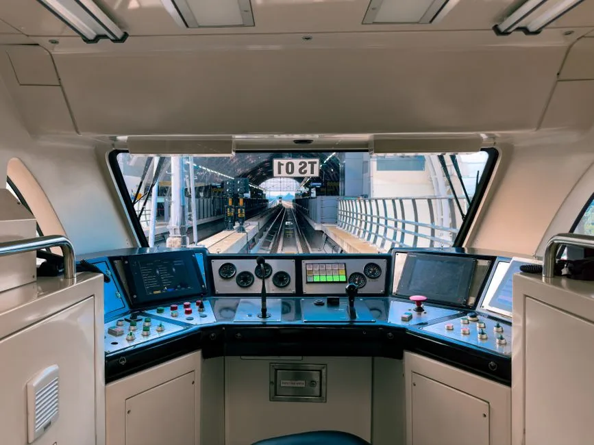 A front-facing view from inside a train or transit vehicle operator’s cab, showing an unbranded control console, screens, gauges, buttons, and tracks through the windshield. No person, passenger seat, or handheld electronic device is visible.
