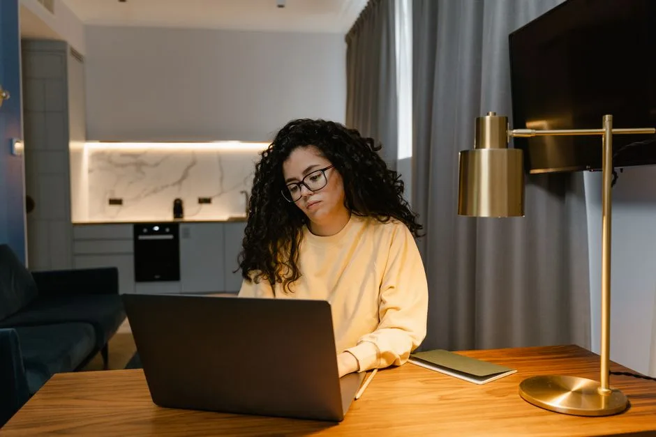 A person wearing glasses sits at a wooden table using an open laptop in a modern living space, with a brass desk lamp, notebook, wall-mounted television, curtains, sofa, and kitchen in the background. No heating product is visibly identifiable.