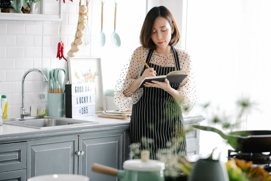 A person wearing an apron writes in a notebook while standing beside a kitchen counter and sink; cookware and generic kitchen items are visible, but no small appliance is clearly being examined.