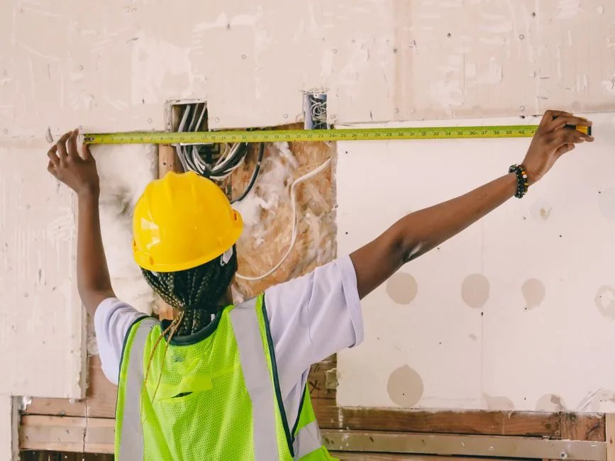 A construction worker wearing a yellow hard hat and high-visibility vest measures an unfinished interior wall with a tape measure; exposed wiring and wall framing are visible.