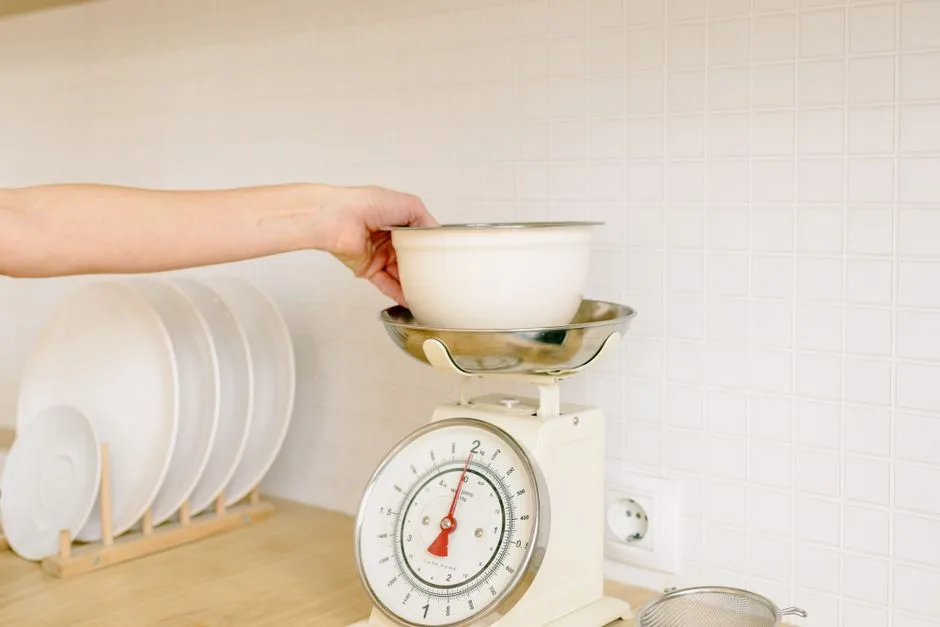 A hand places a plain white bowl on an unbranded analog kitchen scale beside a dish rack in a neutral kitchen.