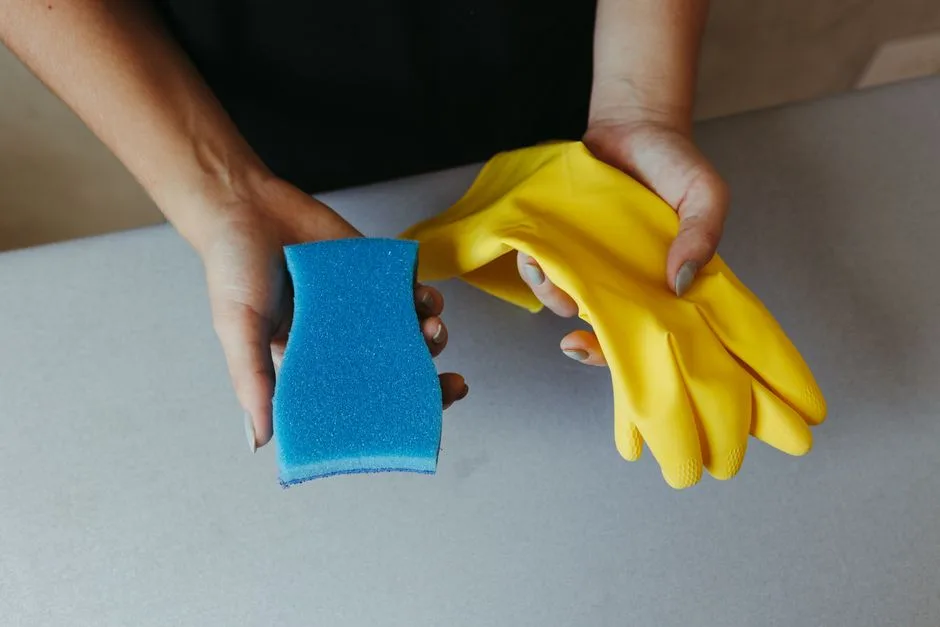 A person holds a blue cleaning sponge in one hand and yellow rubber cleaning gloves in the other above a light gray surface.