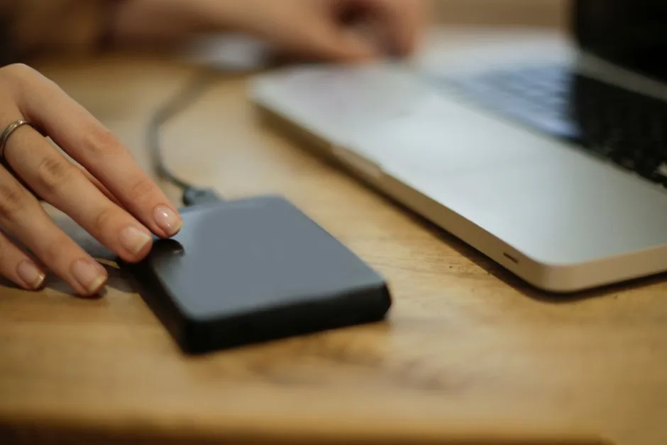 A hand rests on a generic portable external drive connected by a cable beside an open silver laptop on a wooden desk.